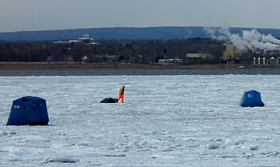 [Person in a yellow jacket and florescent orange pants pulling a sled across the ice.]